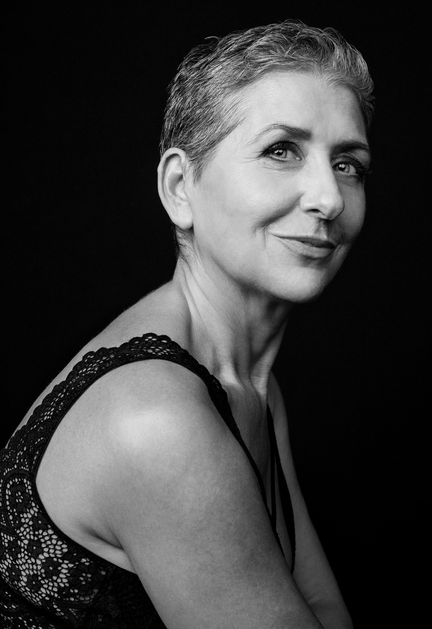 Retrato en blanco y negro de una mujer de pelo cano y corto con sonrisa suave, mirando por encima del hombro sobre fondo negro