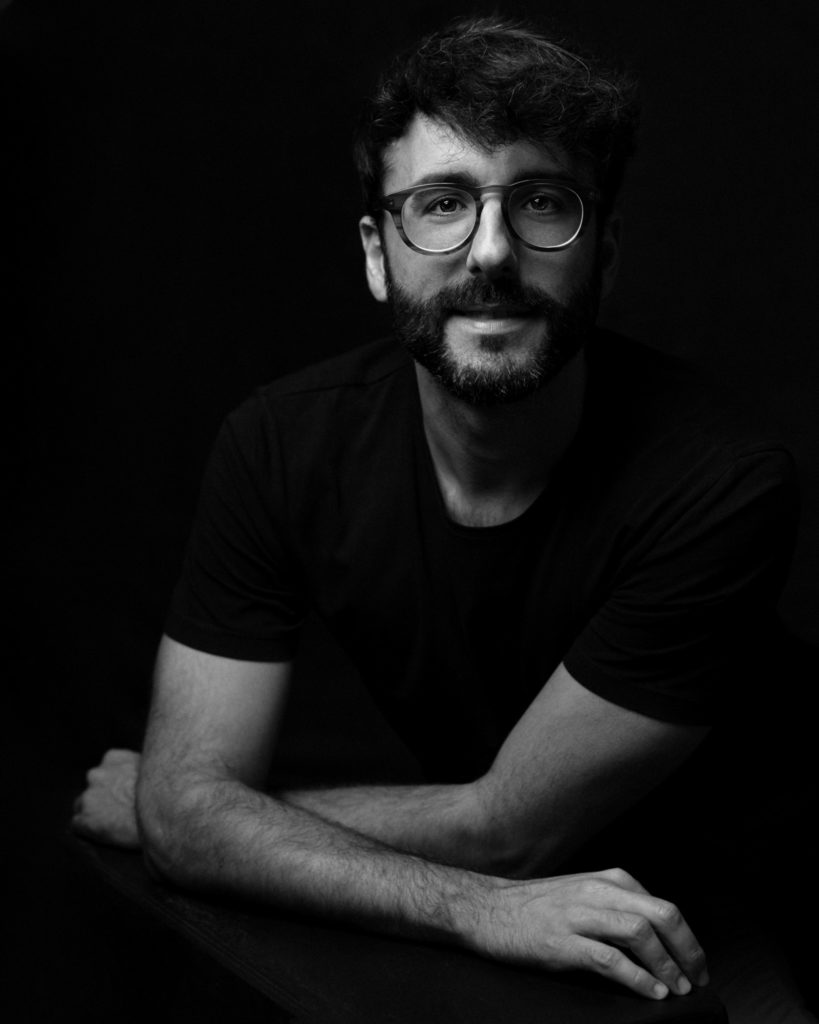 Retrato de estudio de un hombre con gafas y barba, sentado y con los brazos apoyados, sobre fondo negro